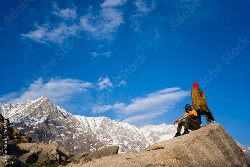 Two tourist young men sitting on rocky cliff and enjoying beautiful view, Triund Trek, Himachal Pradesh, India.