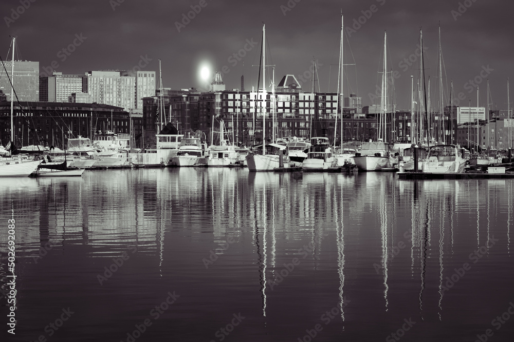 The noir scene with sailboats in front of the city. Sailboat masts without sails reflect dramatically in the calm water.