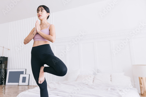 Beautiful happy young Asian woman standing in Vrksasana Posture, doing yoga exercise on purple mat with water bottle, working out in bedroom, indoor workout, Tree Pose.