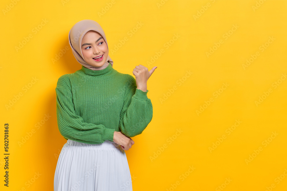 Cheerful beautiful Asian woman in green sweater pointing thumbs aside at copy space isolated over yellow background Cheerful beautiful Asian woman in green sweater pointing thumbs aside at copy space isolated over yellow background