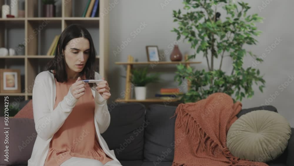 Caucasian young woman sitting alone on couch and feeling shocked with positive unplanned pregnancy test result. Hard decision and unwilling pregnancy concept. Caucasian young woman sitting alone on couch and feeling shocked with positive unplanned pregnancy test result. Hard decision and unwilling pregnancy concept.