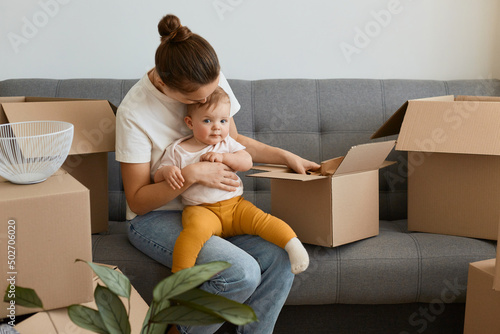 Bilde på lerret Portrait of woman with bun hairstyle wearing white t shirt sitting on sofa and holding her infant baby, unpacking boxes together with her daughter, young family moving into a new apartment