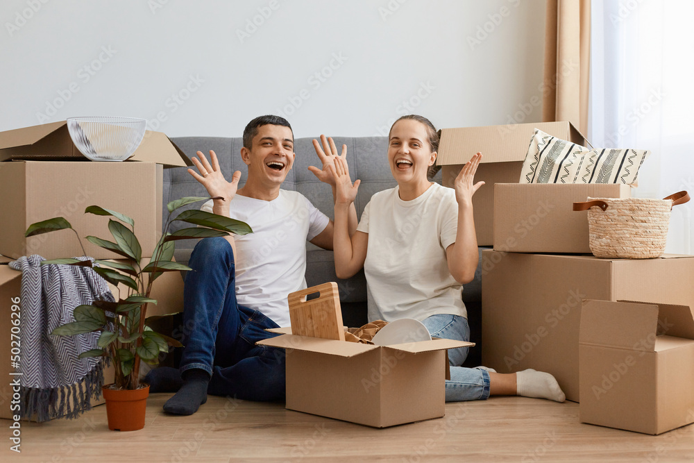 Full length portrait of joyful newlyweds moved in new apartment, sitting on the floor among cardboard boxes, raised hands, rejoicing to rent a house together. Full length portrait of joyful newlyweds moved in new apartment, sitting on the floor among cardboard boxes, raised hands, rejoicing to rent a house together.