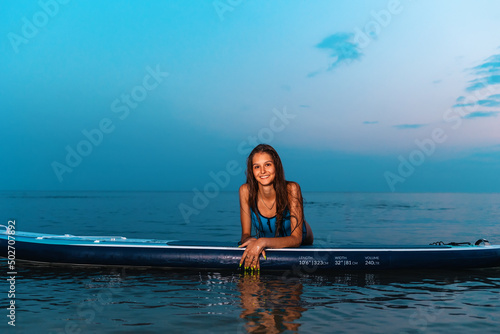 Summer sport. Portrait of young smiling tanned woman poses leaning on a sup board. Copy space. Concept of surfing