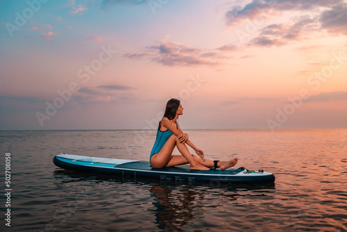 Pretty young woman in blue swimsuit sitting on a sup board. Sunset in the background. Copy space. Concept of water sport and summer activity