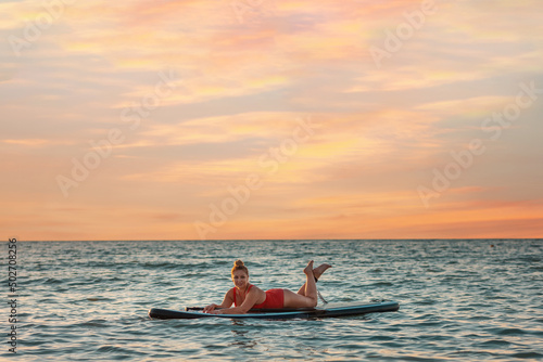 Plus sized beautiful woman in red swimsuit posing lying on a sup board. Copy space. Sunset sky on the background. The concept of sports and summer vacation