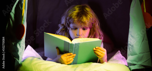 Child reading a book in the dark home. Close up portrait of Boy sitting on in living room watching pictures in story book. Kid doing homework for elementary school. Children study.