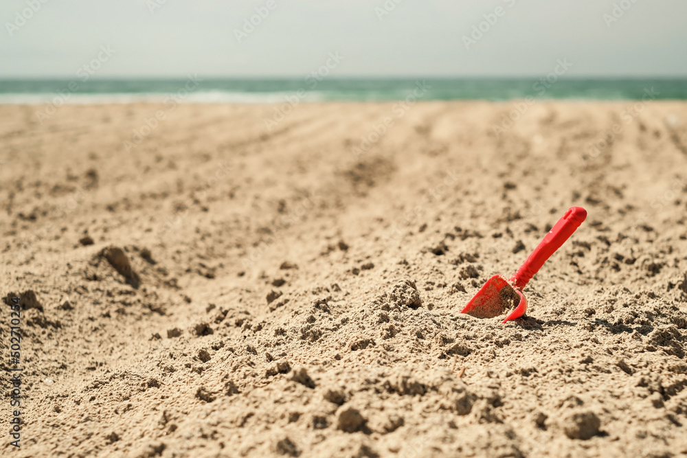 Summer children's beach toy - a red spatula on a sandy beach against the background of the sea and sky. Summer vacation banner idea Summer children's beach toy - a red spatula on a sandy beach against the background of the sea and sky. Summer vacation banner idea