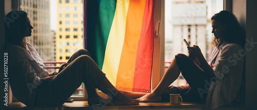 Beautiful female lesbian couple in love sitting near the window with the rainbow flag and using smartphone. Concept LGBT couple at home. photo