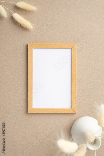 Top view vertical photo of wooden photo frame and white ceramic vase with white lagurus flowers on beige background with copyspace