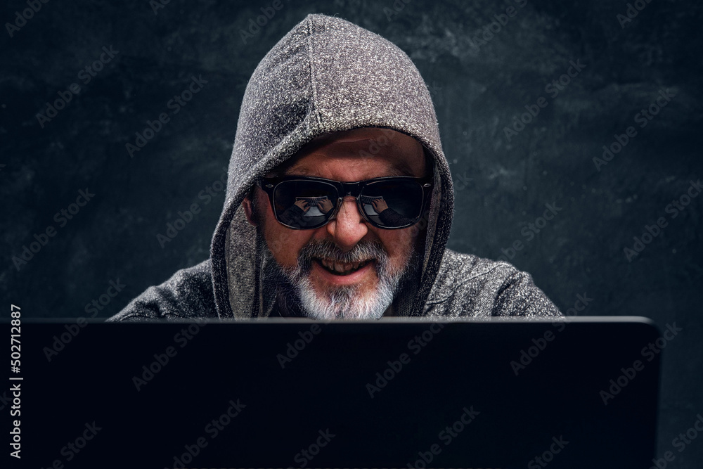 Portrait of old man hacker dressed in sunglasses and hoody using laptop against dark background. Portrait of old man hacker dressed in sunglasses and hoody using laptop against dark background.