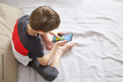 Little boy playing mobile game on smartphone sitting on a sofa, top view. Child leisure at home, video gaming addiction