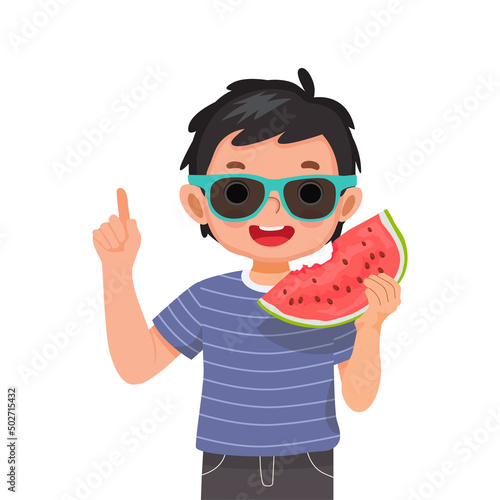 Happy cute little boy with sunglasses eating watermelon pointing finger up on sunny day summer time