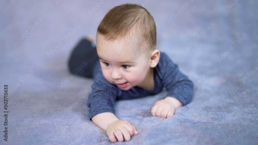 Nice Caucasian baby boy lying on his tummy on big bed. Mom's hand reaches the son and he responds with a cute smile. Grey backdrop in blur. Nice Caucasian baby boy lying on his tummy on big bed. Mom's hand reaches the son and he responds with a cute smile. Grey backdrop in blur.
