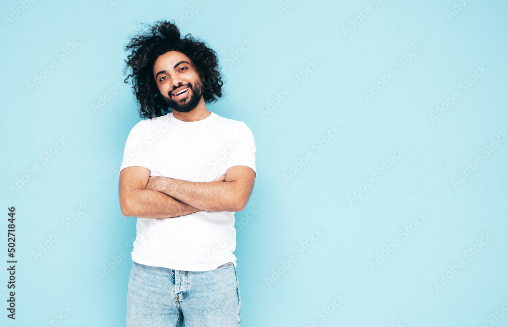 Handsome smiling hipster model.Sexy unshaven Arabian man dressed in white summer t-shirt and jeans clothes. Fashion male with long curly hairstyle posing near blue wall in studio Handsome smiling hipster model.Sexy unshaven Arabian man dressed in white summer t-shirt and jeans clothes. Fashion male with long curly hairstyle posing near blue wall in studio