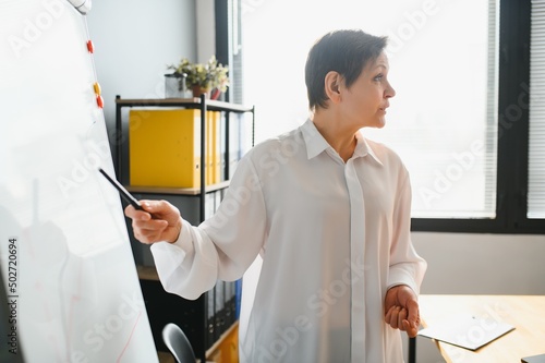 Smiling old mature female coach manager trainer pointing on whiteboard give business flip chart presentation, happy senior adult teacher mentor speaker presenter conduct conference workshop concept
