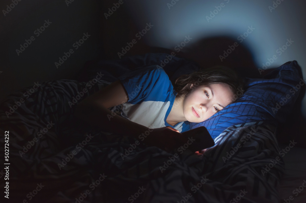 The girl lies at home in the evening on the bed with a phone in the dark close-up. defocus The girl lies at home in the evening on the bed with a phone in the dark close-up. defocus
