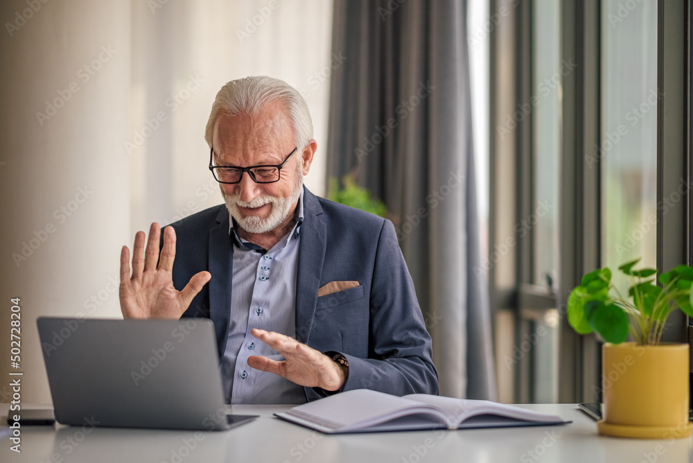 Elderly businessman greeting while video conferencing on laptop at office desk Elderly businessman greeting while video conferencing on laptop at office desk