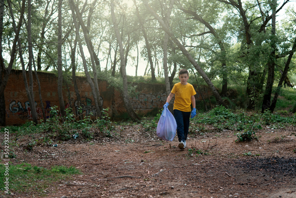 Young boy with a yellow t-shirt and blue gloves walking with a garbage and recycling bag in his hand while observing the river environment to be ecological Young boy with a yellow t-shirt and blue gloves walking with a garbage and recycling bag in his hand while observing the river environment to be ecological