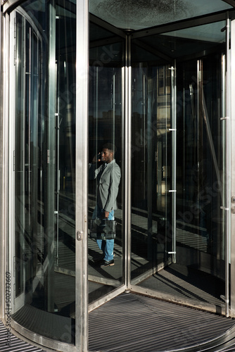 African American businessman standing behind glass revolving doors in business center and talking on the phone
