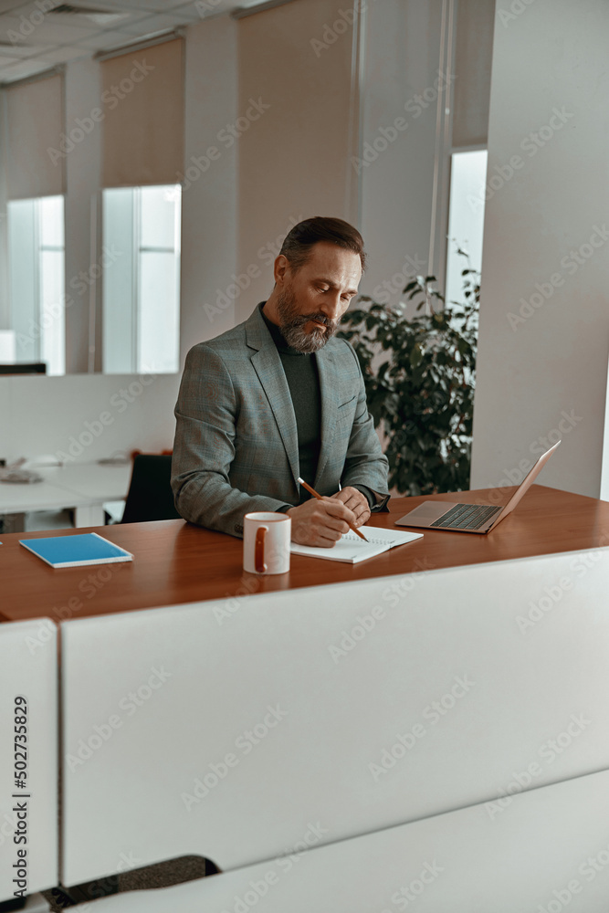 Bearded businessman in casual clothes making notes in notepad while sitting in his office Bearded businessman in casual clothes making notes in notepad while sitting in his office
