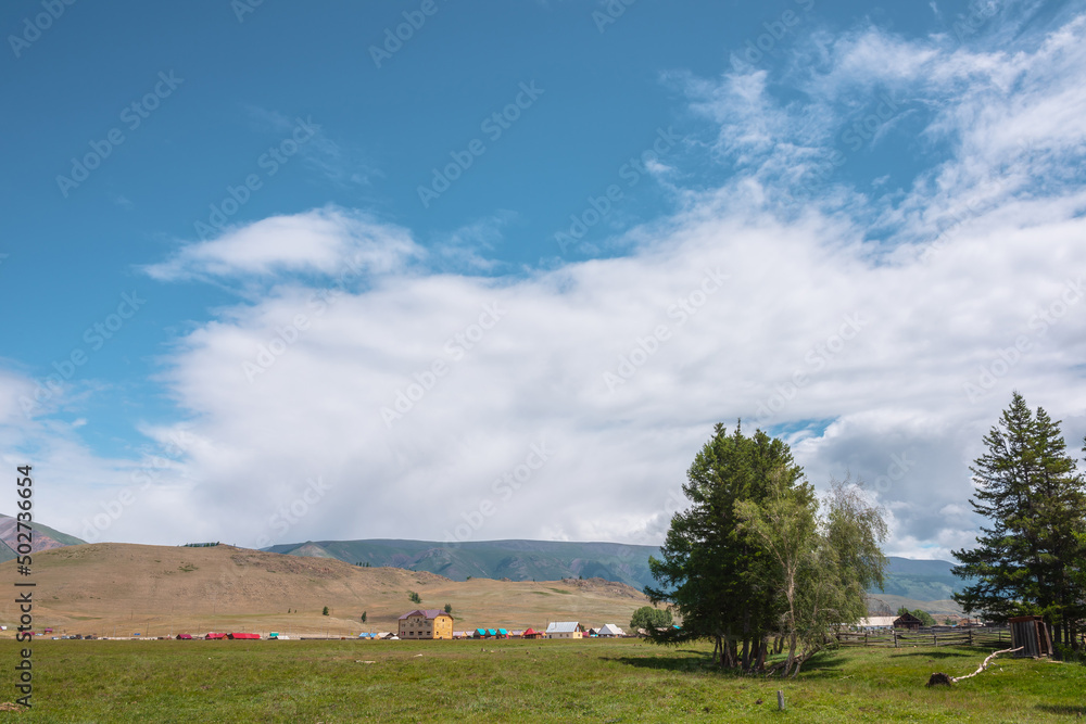 Obraz premium Dramatic alpine view from trees to small houses in sunlit green meadow against high mountain range under clouds in blue sky. Scenic mountain landscape at changeable weather. Village in high mountains.