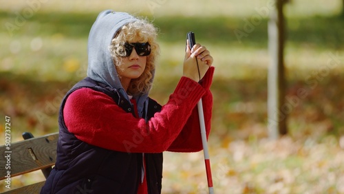 Outdoor portrait of a sad blonde caucasian disabled girl with a hood on her head and sunglasses on her eyes, holding her hands on a white crane. High quality photo