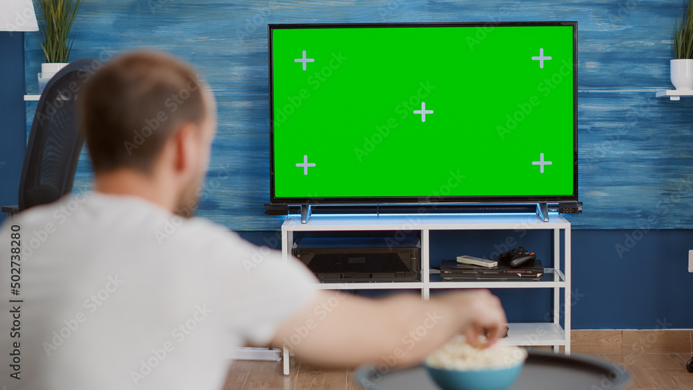 Over shoulder view of sports fan watching game on green screen tv mockup encouraging favourite team sitting on couch. Man sport supporter looking at television with chroma key display in living room. Over shoulder view of sports fan watching game on green screen tv mockup encouraging favourite team sitting on couch. Man sport supporter looking at television with chroma key display in living room.