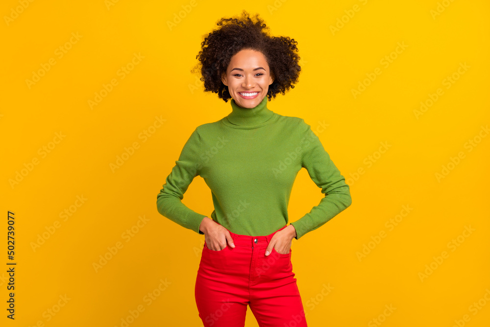Portrait of attractive cheerful wavy-haired girl holding hands in pockets isolated over bright yellow color background