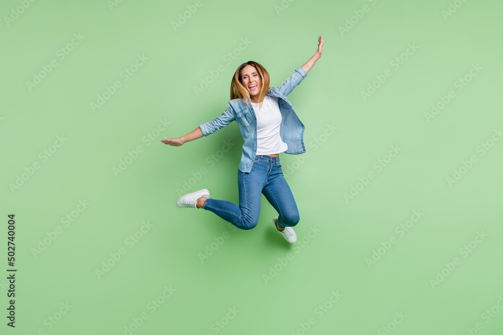 Full size photo of mature lady jump fly wear shirt jeans sneakers isolated on green background Full size photo of mature lady jump fly wear shirt jeans sneakers isolated on green background
