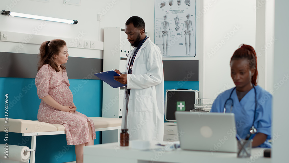 Fototapeta premium African american medic consulting woman with pregnancy belly, taking notes on checkup files. Male obstetrician attending examination appointment with pregnant patient, giving advice.