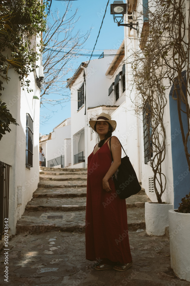 Fototapeta premium young pregnant woman with red dress and hat walking in traditional spanish town with white buildings and clear blue sky on a sunny day, with traditional spanish architecture