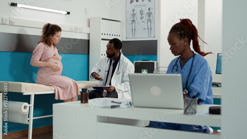 Male doctor meeting with patient expecting child in cabinet, attending maternity appointment and taking notes on checkup papers. Pregnant woman doing consultation with physician.