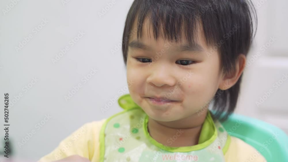 Asian boy sits in a high chair drinking nectar with a spoon and looking at the tablet with a smiling face in the house. Asian boy sits in a high chair drinking nectar with a spoon and looking at the tablet with a smiling face in the house.