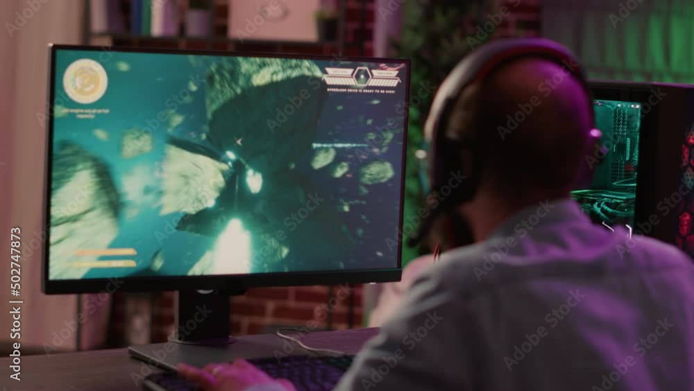 Overs shoulder view of gamer streaming fast paced space shooter action game on gaming pc. Man playing multiplayer online tournament talking to team on headset while explaining gameplay to subscribers. Overs shoulder view of gamer streaming fast paced space shooter action game on gaming pc. Man playing multiplayer online tournament talking to team on headset while explaining gameplay to subscribers.