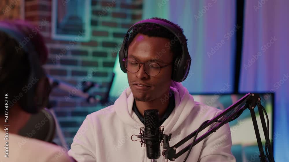 Internet podcaster reading question from laptop screen talking using microphone to influencer guest in home recording studio. African american online radio host in terview with caucasian blogger. Internet podcaster reading question from laptop screen talking using microphone to influencer guest in home recording studio. African american online radio host in terview with caucasian blogger.