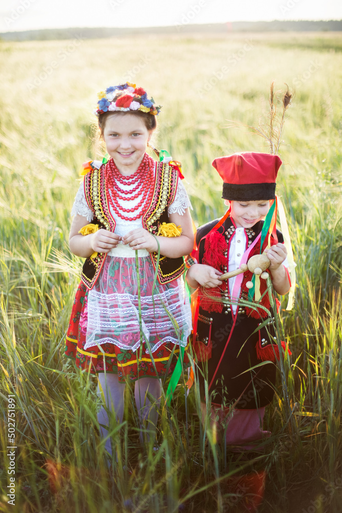 Dzieci w tradycyjnych polskich krakowskich strojach spacerują po polu obsianym zbożem w ciepłych letnich promieniach słonca - obrazy, fototapety, plakaty 