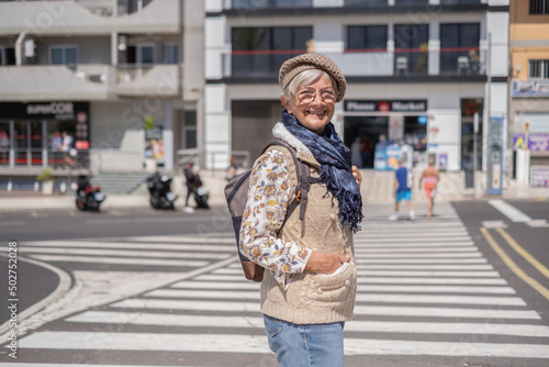 Happy senior woman traveler in sunny city centre expressing positivity, good mood, holding backpack. Attractive elderly lady looking at camera smiling