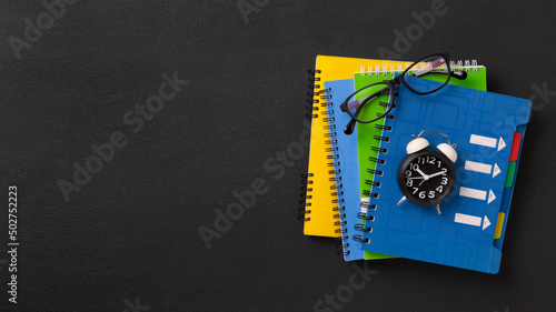 A stack of colored school notebooks and an alarm clock on a school chalkboard background. Back to school concept, banner