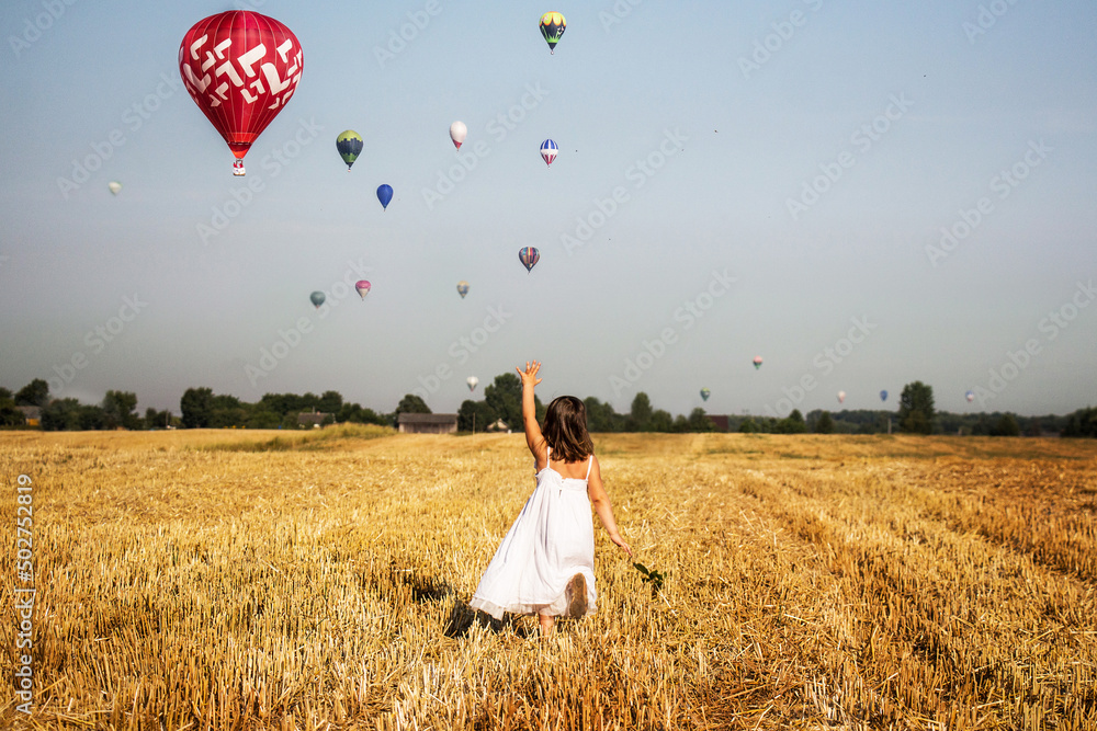 Dziewczynka biegnie po polu i macha do balonów na gorące powietrze, lot balonem nad polami w środku lata - obrazy, fototapety, plakaty 