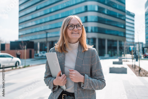 European 30s businesswoman in stylish clothes with a laptop walking outdoors near the business center