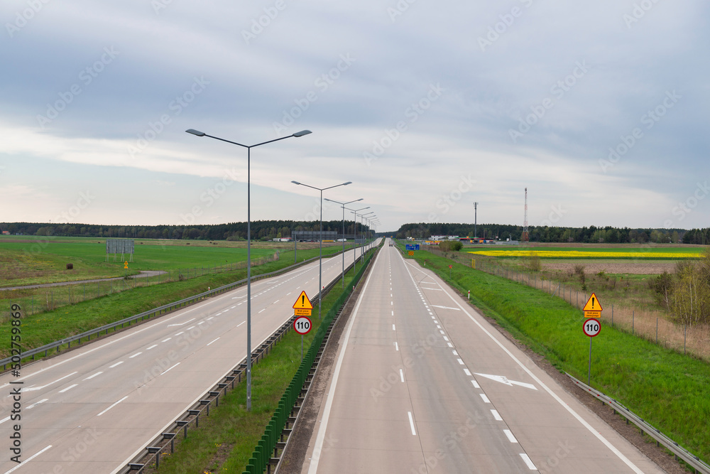 Słoneczny dzień. Niebo lekko zachmurzone, równina pokryta polami uprawnymi i łąkami. Na równinie jest betonowa, dwupasmowa droga. Widać jadące po niej samochody. Słoneczny dzień. Niebo lekko zachmurzone, równina pokryta polami uprawnymi i łąkami. Na równinie jest betonowa, dwupasmowa droga. Widać jadące po niej samochody.