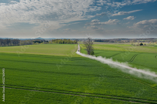 Równina pokryta łąkami i polami sfotografowana z drona. Droga polna przebiegająca przez pola i łąki widziana z wysokości. Zdjęcie z drona. W oddali widać zabudowania pobliskiej wioski.