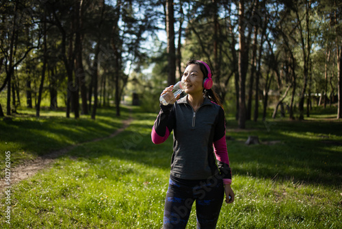 Young Asian woman in sportswear drinking water from a bottle while jogging at city public park in the morning. Healthy female athlete enjoy outdoor lifestyle sport training workout running exercise 