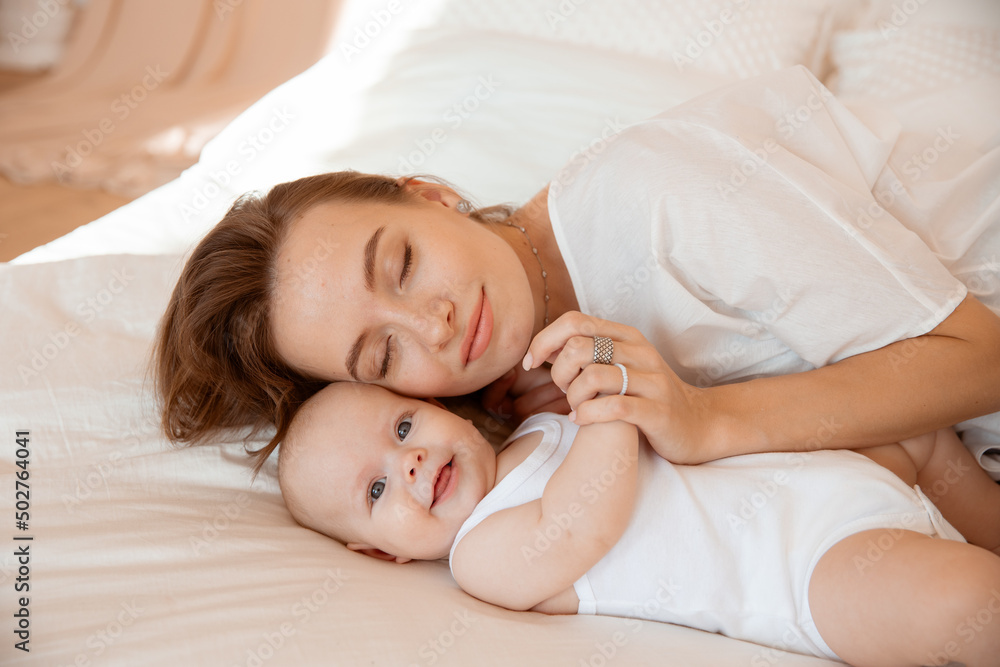 Happy young mother with baby at home in white room, mom and baby are lying, kissing the baby at home on the bed in the bedroom Happy young mother with baby at home in white room, mom and baby are lying, kissing the baby at home on the bed in the bedroom