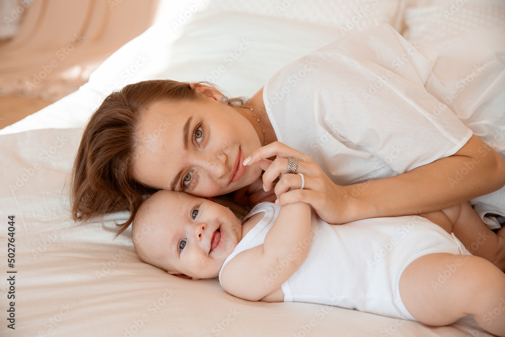 Happy young mother with baby at home in white room, mom and baby are lying, kissing the baby at home on the bed in the bedroom Happy young mother with baby at home in white room, mom and baby are lying, kissing the baby at home on the bed in the bedroom