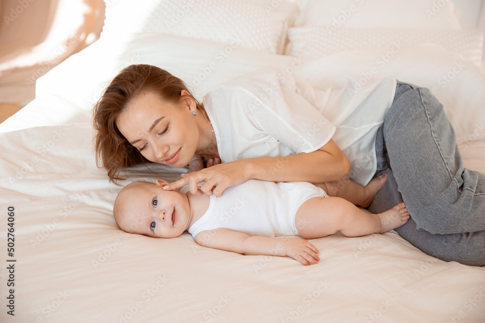 Happy young mother with baby at home in white room, mom and baby are lying, kissing the baby at home on the bed in the bedroom Happy young mother with baby at home in white room, mom and baby are lying, kissing the baby at home on the bed in the bedroom