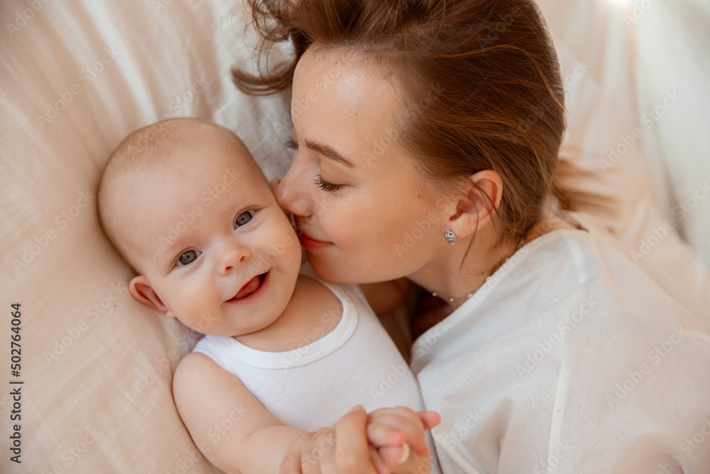 Happy young mother with baby at home in white room, mom and baby are lying, kissing the baby at home on the bed in the bedroom Happy young mother with baby at home in white room, mom and baby are lying, kissing the baby at home on the bed in the bedroom