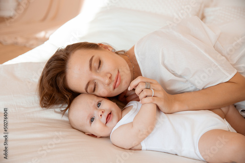 Happy young mother with baby at home in white room, mom and baby are lying, kissing the baby at home on the bed in the bedroom