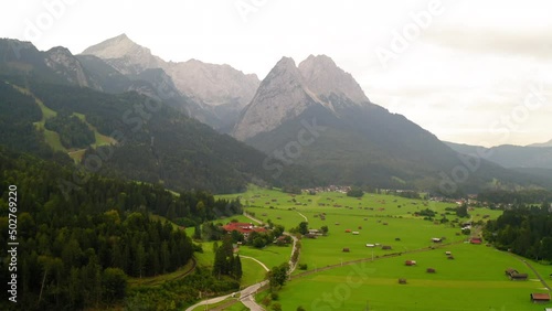Aerial Beautiful Shot Of Town By Forest On Mountains, Drone Flying Forward Over Paragliding Landing Field - Garmisch-Partenkirchen, Germany